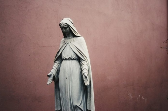Estátua de Nossa Senhora com as mãos estendidas diante de uma parede cor de vinho
